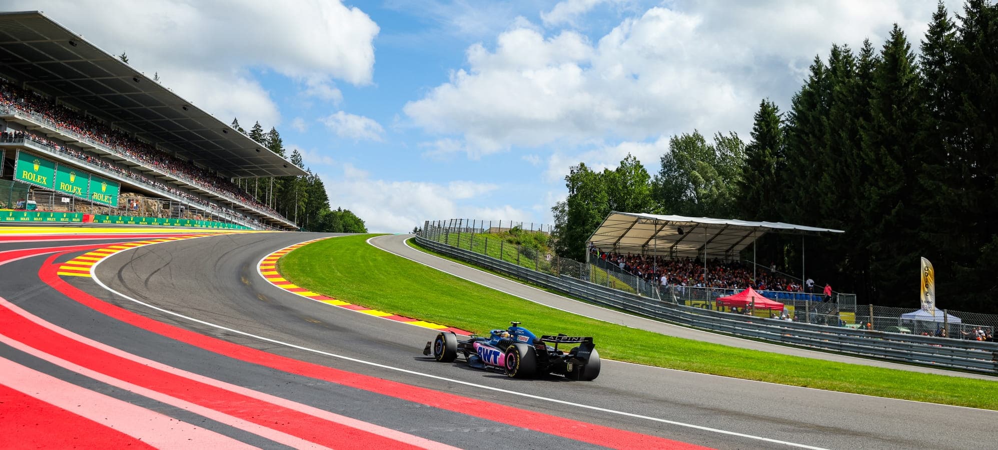 A Formula 1 car climbs through Eau Rouge and Raidillon at Spa-Francorchamps beneath a bright Ardennes sky
