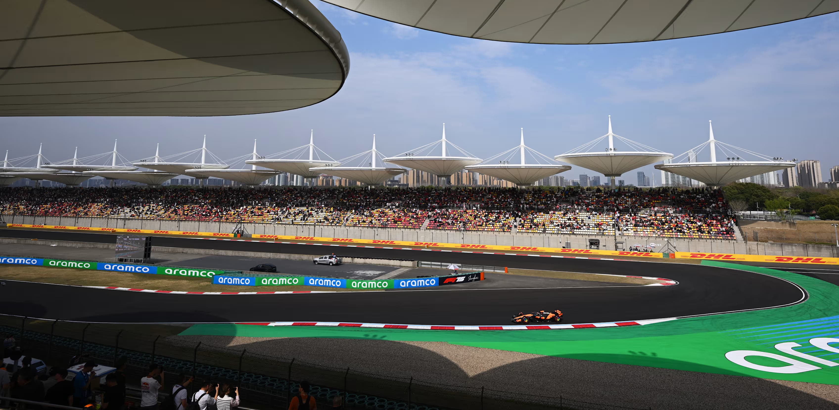 An orange Formula 1 car rounds the final corner at Shanghai International Circuit beneath the circuit's distinctive grandstand canopies
