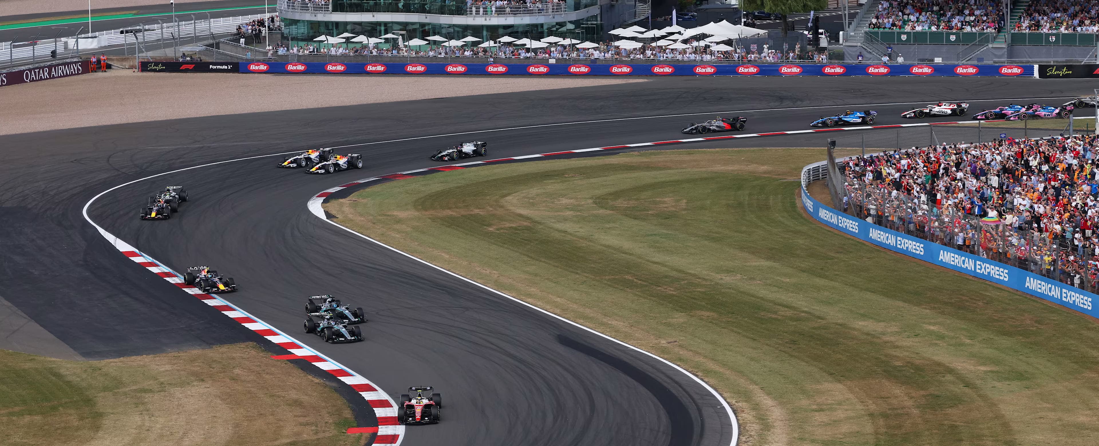 A line of Formula 1 cars flows through a broad Silverstone corner beside full grandstands
