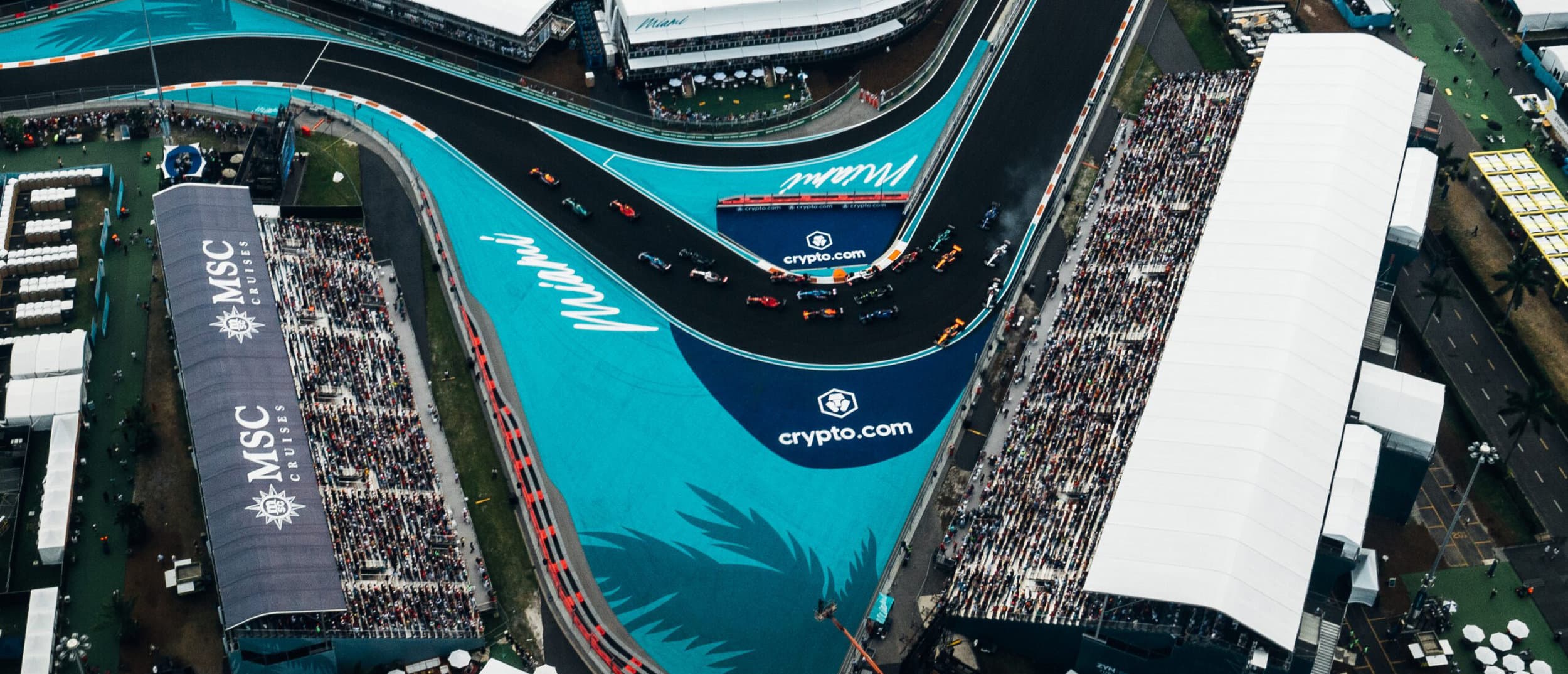 An aerial view of Formula 1 cars racing through the turquoise-lined Miami International Autodrome with packed grandstands on both sides